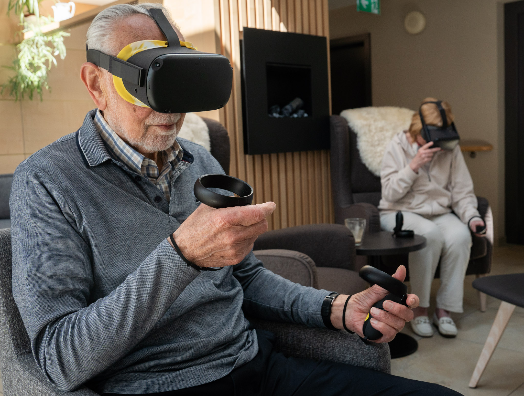 Un homme et une femme âgés sont assis sur des chaises confortables dans un salon et s'entraînent avec des lunettes VR et des manettes.